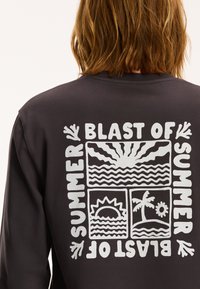 Crna majica dugih rukava s bijelim grafičkim dizajnom na leđima koji sadrži motive sunca, vala i palme, te natpis "BLAST OF SUMMER."
