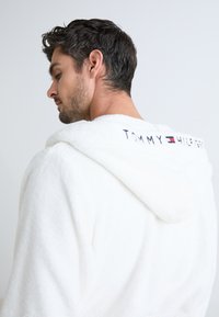 Accappatoio bianco in peluche con cappuccio, dotato di logo ricamato "TOMMY HILFIGER" sulla schiena. Tessuto morbido e vestibilità comoda.