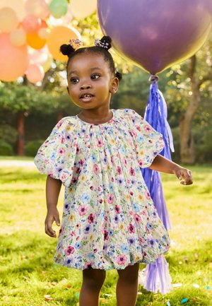 Junges Mädchen mit Blumen-Kleid und Duttfrisur steht draußen neben einem großen violetten Ballon und bunten Dekorationen in einem sonnigen Garten.