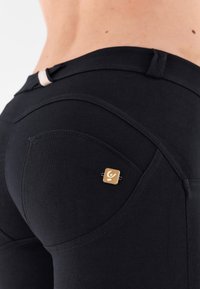 Czarne dopasowane spodnie o bezszwowym designie, z małym złotym logo i detalami przeszycia na tylnej kieszeni. Gładka tekstura.