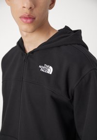 Czarna bluza zapinana na zamek, wykonana z miękkiego materiału, z kieszenią typu kangur oraz białym logo na lewej piersi, posiadająca usztywniony kaptur.