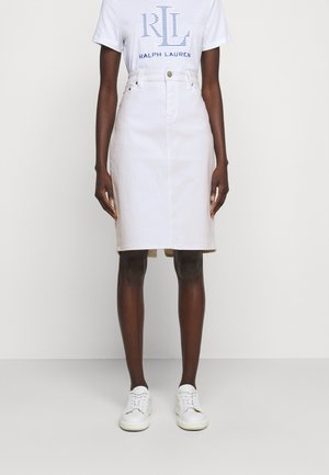 Person iført en hvid Ralph Lauren logo T-shirt, hvid high-waisted knælange nederdel og hvide sneakers, stående foran en ensfarvet baggrund.