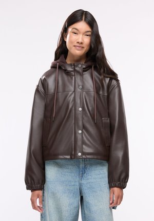 Giovane donna con lunghi capelli scuri indossa una giacca marrone in pelle con bottoni e cappuccio e jeans azzurri davanti a uno sfondo bianco semplice.