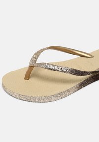 Flip-flopuri aurii cu sclipici, având un toc din cauciuc texturat bej, bretele subțiri și accente metalice, etichetate "havaianas".