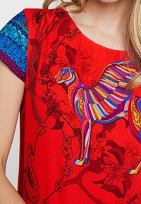 Czerwony top z krótkim rękawem z kolorowym wzorem kota z skrzydłami. Ozdobiony niebieskimi wzorami kwiatowymi na rękawach oraz misternymi detalami kwiatowymi.