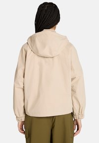 Chaqueta beige con capucha, puños elásticos y corte relajado. Presenta un sutil logo en la parte posterior. Tejido ligero y textura suave.
