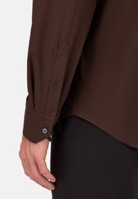 Ruda ilga rankovė marškiniai su glotnia tekstūra, turi sagų apykakles ir detalią siuvinėjimą. Riba šiek tiek išlenkta.