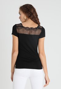 Schwarzes T-Shirt mit kurzen Ärmeln, ausgestattet mit einem transparenten Spitzenpanel am Rücken und einer figurbetonten Silhouette. Weiße Hose sichtbar an der Taille.