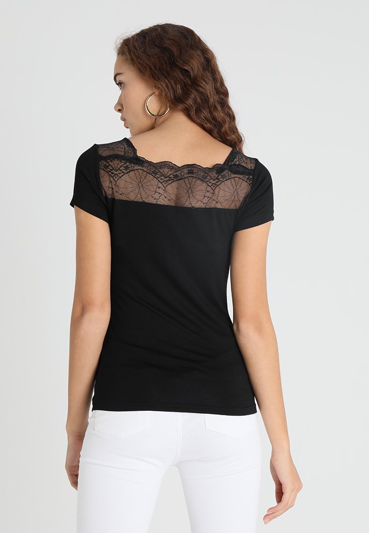 Schwarzes T-Shirt mit kurzen Ärmeln, ausgestattet mit einem transparenten Spitzenpanel am Rücken und einer figurbetonten Silhouette. Weiße Hose sichtbar an der Taille.