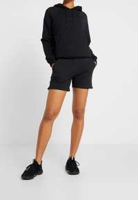Hoodie noir avec poche avant et cordon de serrage, associé à un short noir. Le modèle porte des chaussures de sport noires et se tient devant un fond blanc.