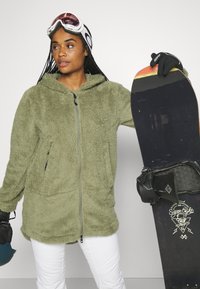 Olivgrüne Fleecejacke mit Reißverschluss, langen Ärmeln und Seitentaschen. Das Model hält ein Snowboard, trägt weiße Hosen und Skibrille.