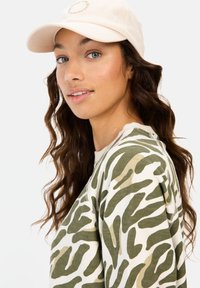 Beige baseball cap med et logo, parret med en langærmet top med et olivengrønt og cremet abstrakt mønster. Bølget brunt hår synligt.