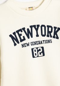 Kremowy sweatshirt wykonany z bawełny, z granatowym napisem "NOWY JORK" oraz "NOWE POKOLENIA" z numerem "82" poniżej. Żebrowany dekolt.
