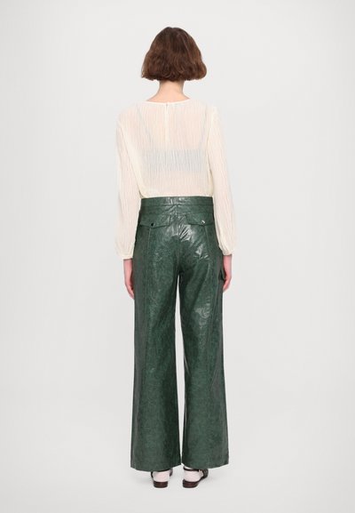 Crème semi-transparante top met lange mouwen en een sleuf aan de achterkant, gecombineerd met hooggetailleerde groene neplederen wijde broek.