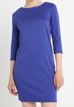 Vrouw met een eenvoudige blauwe jurk tot op de knieën en driekwart mouwen, staand tegen een witte achtergrond.