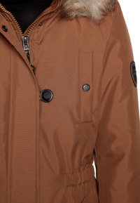 Veste d'hiver marron avec fermeture éclair, bouton noir, poche à pression, taille élastique et col bordé de fourrure, portée sur un pull noir.