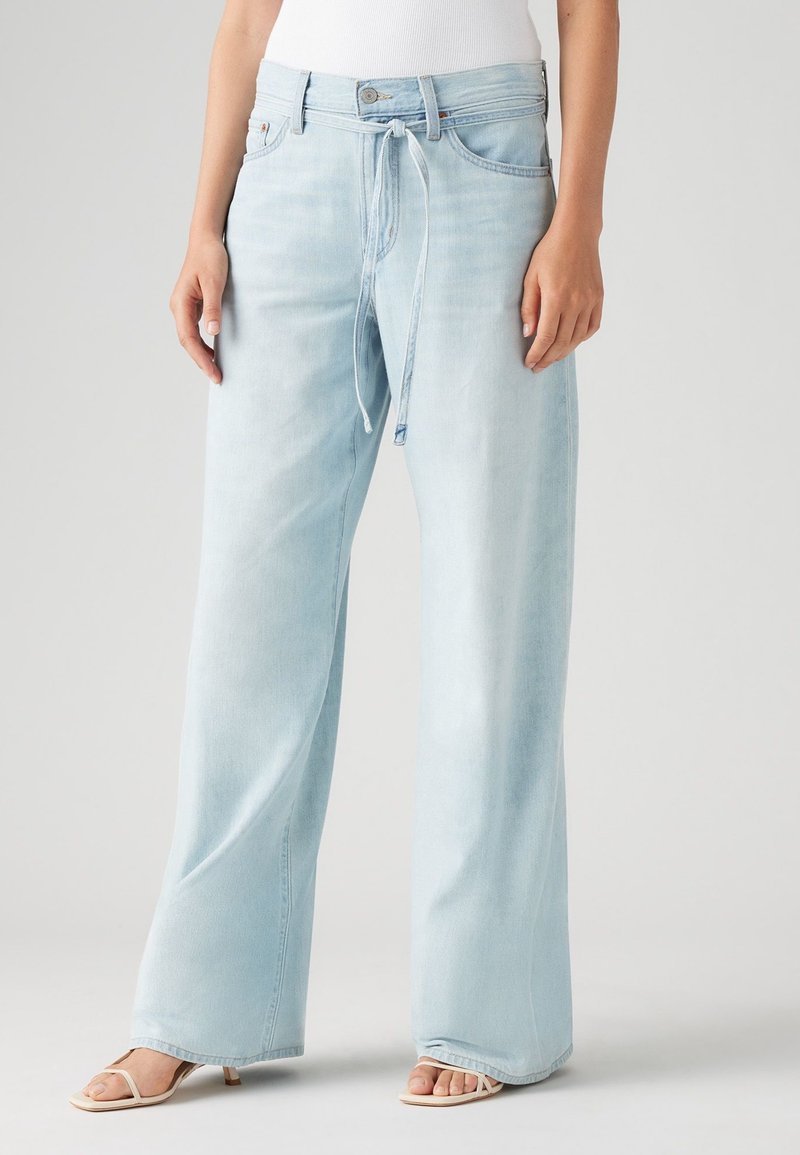 Personne portant un jean bleu clair à jambes larges avec un cordon noué à la taille et des sandales blanches à talons.