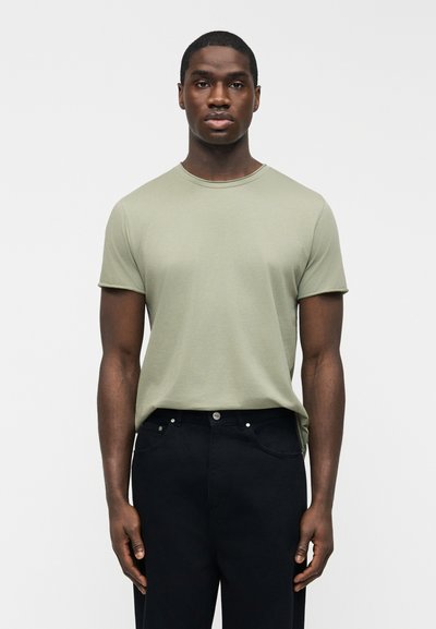Ung man med kort hår bär en ljusgrön kortärmad T-shirt och svarta byxor, stående mot en slät vit bakgrund.
