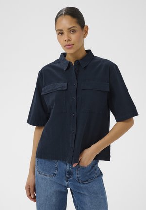 Kobieta ubrana w granatową koszulę z krótkim rękawem na guziki z kieszeniami na piersi oraz niebieskie dżinsy, stojąca z jedną ręką w kieszeni na jednolitym tle.