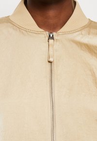 Chaqueta bomber beige con cuello acanalado, que presenta un cierre frontal con cremallera y un acabado ligeramente texturizado. Diseño minimalista y color neutro.