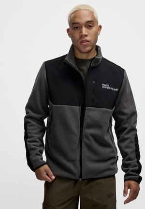 Jonge man met kort blond haar die een zwart-grijs fleece jack met rits draagt met de tekst "HALO" op de borst, staand tegen een effen achtergrond.