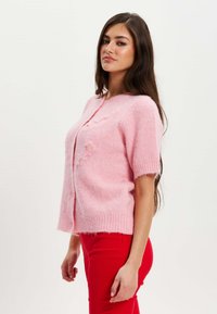 Pink short-sleeve cardigan with button closure, featuring a soft, fuzzy texture and embroidered details. Paired with red fitted pants.