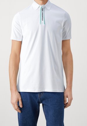 Weißes Polohemd mit kurzen Ärmeln und Kragen, grün eingefasste Knopfleiste, mit dem Schriftzug „KARL LAGERFELD“, getragen zu blauen Jeans an einem stehenden männlichen Oberkörper.