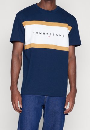 Homme portant un t-shirt Tommy Jeans bleu marine avec des rayures horizontales blanches et moutarde, associé à un jean bleu.