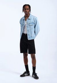 Chaqueta de mezclilla azul claro, camiseta de tirantes blanca, pantalones cortos negros, zapatillas negras con rayas blancas y calcetines negros; pose de pie frente a un fondo liso.