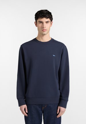 Mladi moški s temnimi kodrastimi lasmi, oblečen v mornarsko moder pulover z majhno svetlo modro pasjo nalepko na prsih in temne kavbojke.