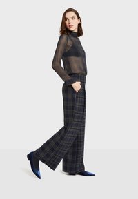 Průhledný černý top s dlouhými rukávy a vysokým límcem, který je spárován s tmavě kostkovanými kalhotami s širokými nohavicemi, obsahujícími zelené a béžové pruhy. Modré semišové boty.