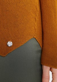 Oranssi neulepaita, jossa on teksturoitu kuvio ja hopeinen logotagi. Helmassa on lievä V-muoto, jonka alla on vihreä kangas.