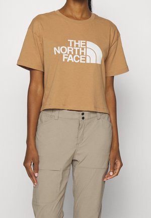 Henkilö pukeutuneena lyhytresuiseen vaaleanruskeaan t-paitaan, jonka edessä on "The North Face" -logo, ja vaaleanbeigeihin housuihin, seisoo yksinkertaisen valkoisen taustan edessä.