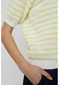 Ruka osoby oblečené v bílo-žlutém pruhovaném krátkém rukávu s rukou odloženou v kapse z tmavého denimu.