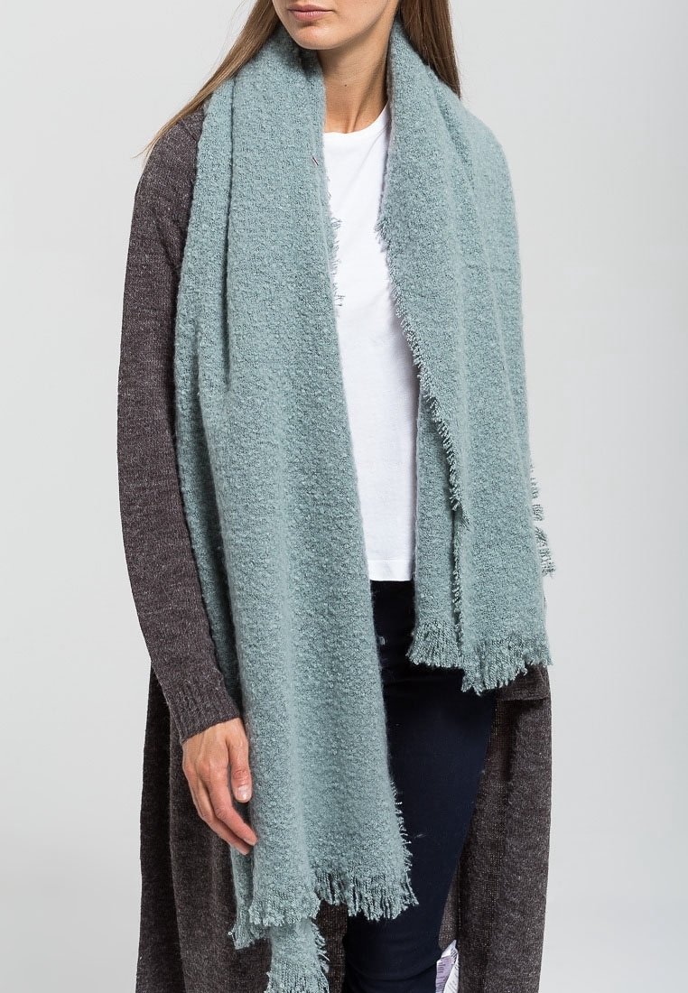 Femme portant une écharpe douce, bleu clair, texturée avec des bords à franges, par-dessus un haut blanc et un cardigan en maille foncé.