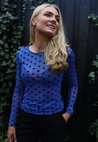 Kék hosszú ujjú felső áttetsző anyagból, apró fekete szívmintákkal díszítve. Testhez simuló kialakítás, sima textúrával és kerek nyakkivágással.