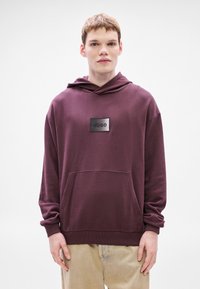 Felpa con cappuccio color amaranto realizzata in tessuto morbido, con tasca frontale e patch logo centrale. Maniche lunghe con polsini e orlo a costine.