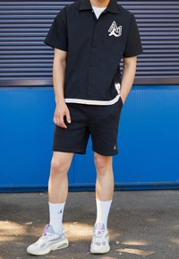 Svart kortärmad skjorta med krage, vit kant och logotyp, kombinerad med svarta shorts. Bärs med vita strumpor och grå sneakers med lila detaljer.