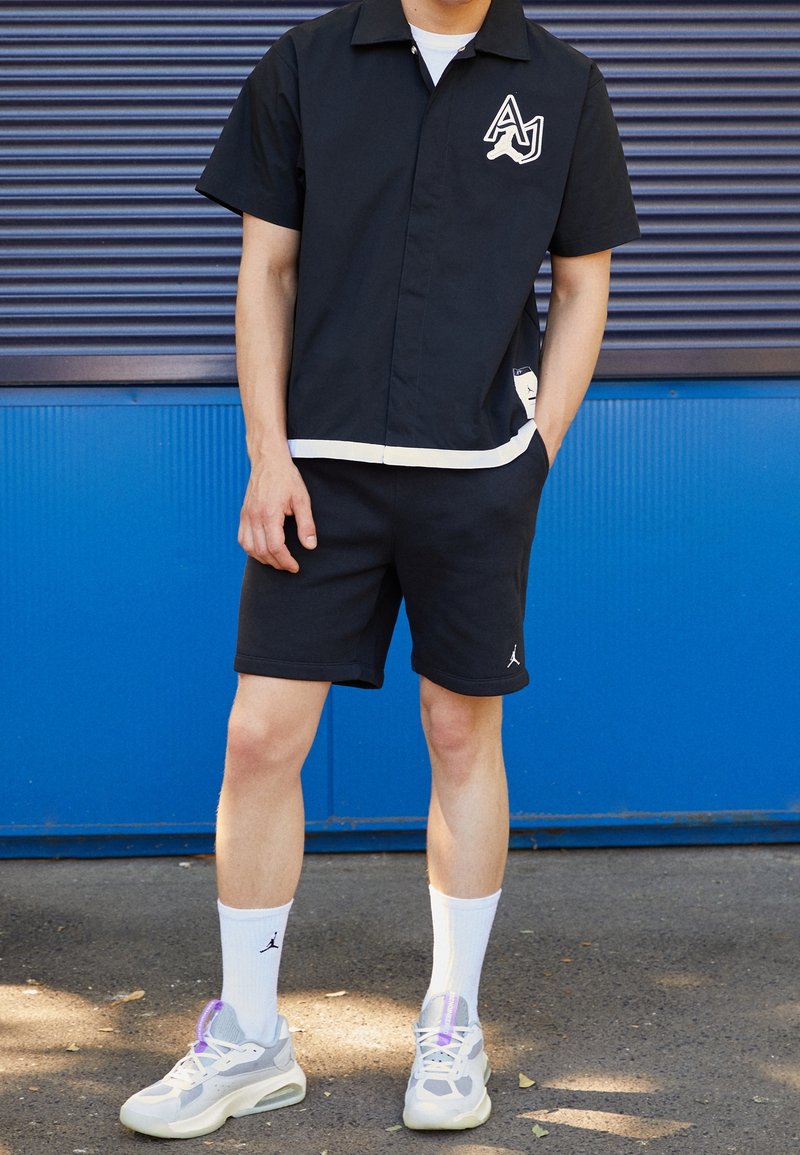 Svart kortärmad skjorta med krage, vit kant och logotyp, kombinerad med svarta shorts. Bärs med vita strumpor och grå sneakers med lila detaljer.