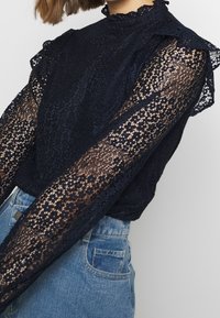 Námořnická blůza s vysokým límcem, krajkovými detaily, průhlednými dlouhými rukávy, volánkovými akcenty a přiléhavým střihem, zkombinovaná s modrými džíny.