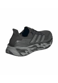 Chaussure de sport noire avec une tige texturée, arborant trois rayures grises distinctives, et une semelle extérieure moulée avec un motif en vagues.