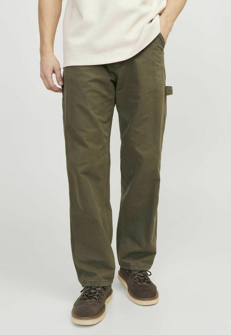 Homme portant un pantalon large vert olive et des chaussures à lacets marron foncé, debout avec une main dans la poche.