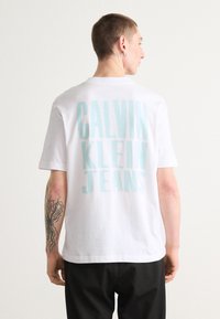 Bílá bavlněná tričko s velkým světle modrým nápisem "CALVIN KLEIN JEANS" na zadní straně. Krátké rukávy a standardní střih.