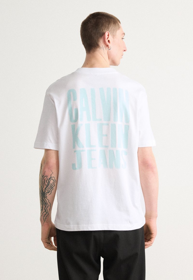 Bílá bavlněná tričko s velkým světle modrým nápisem "CALVIN KLEIN JEANS" na zadní straně. Krátké rukávy a standardní střih.