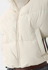 Light beige puffer jacket with a zip front, side pockets, and elastic cuffs. Smooth texture, slightly cropped design with horizontal stitching.