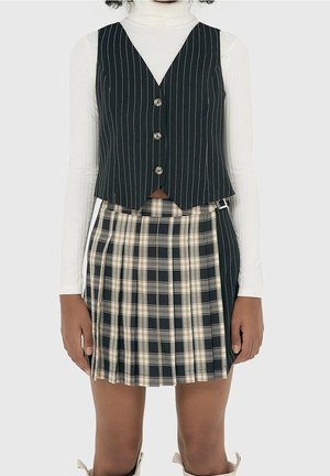 Femme portant un gilet noir à fines rayures boutonné sur un col roulé blanc à manches longues et une jupe plissée à carreaux noire, beige et blanche.