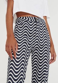 Persona vestida con pantalones de cintura alta de patrón ondulado en blanco y negro, y una blusa corta blanca, con las manos descansando a los lados contra un fondo liso.