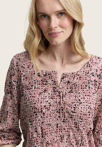 Lässige pinke Bluse mit einem strukturierten Muster aus schwarz-weißen Punkten, ausgestattet mit einem V-Ausschnitt und Knopfakzenten auf der Vorderseite.