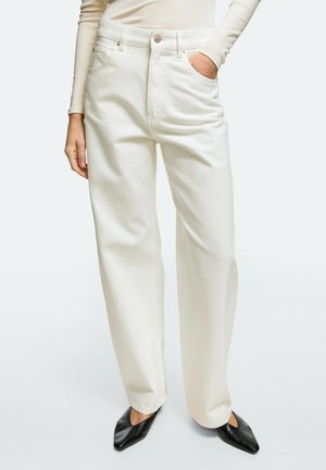 Donna che indossa pantaloni bianchi a vita alta a gamba larga, top beige a maniche lunghe, con una mano in tasca e scarpe nere a punta.