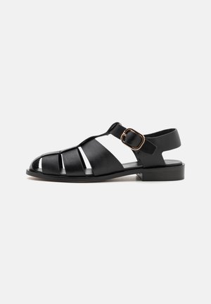 Black leather sandals with a closed toe, open sides, and a buckle strap. Flat heel and a smooth, shiny texture.